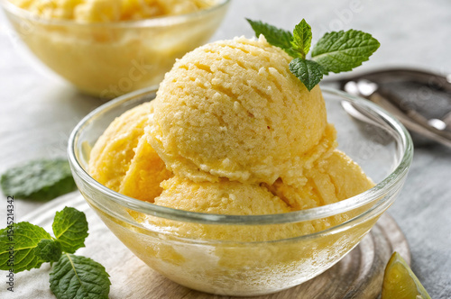 shows a bowl of yellow sorbet with a mint leaf on top, placed on a wooden board with a napkin under it. Another bowl of sorbet and a lime are visible in the background.