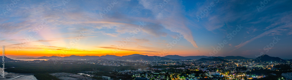 Obraz premium A stunning panoramic view of a coastal city at sunset at Phuket city, showcasing vibrant hues of orange and blue in the sky, with mountains, and urban lights creating a harmonious blend of natural