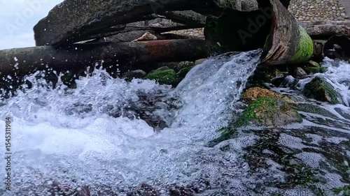 A stream of clean fresh water with foam and air bubbles flows out of an old iron pipe and washes the stones with green algae