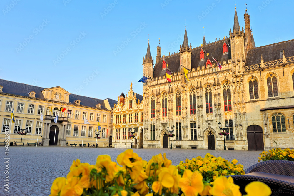 Fototapeta premium Bruges City Hall under the warm glow of sunset with yellow flowers in the foreground, Belgium