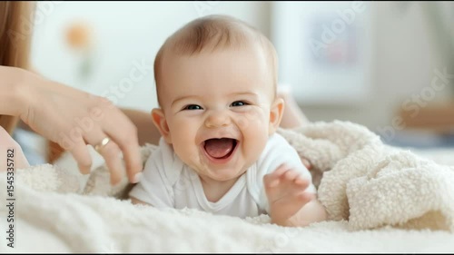 Adorable laughing baby wearing a white onesie lies on a soft blanket on bed. Mother hand is gently supporting newborn baby