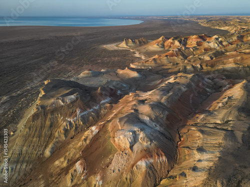 The video moves along curved, stratified slopes of Aktumsuk, exposing bands of red, white, and orange. The vast, dry steppe meets the tranquil blue water of the receded Aral Sea in the distance