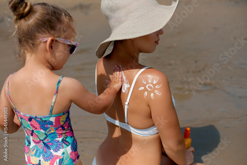 A young child helps apply sunscreen to a seated woman on a sunny beach. The image captures a moment of care and cautious protection against the sun in a colorful summer sett.