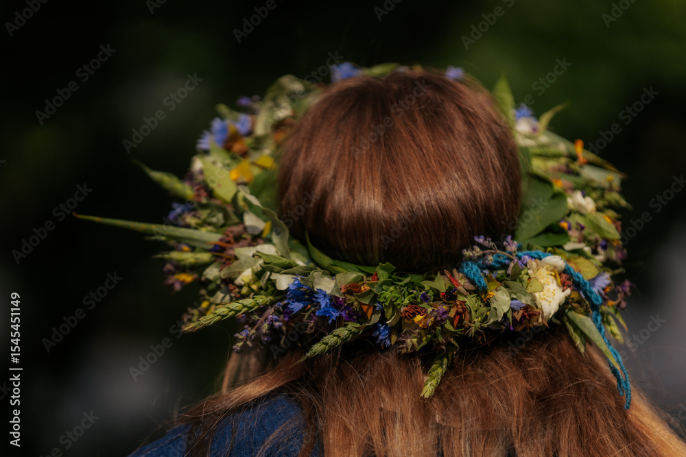 Naklejka premium Close-up of a flower crown made of colorful wildflowers and greenery on a woman's brown hair, viewed from behind during a traditional outdoor celebration.