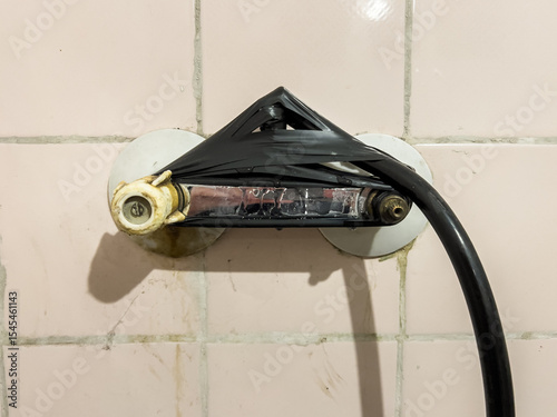 Provisionally repaired shower holder, wrapped with black insulating tape, on an old, pink, dirty tile wall, showing improvisation and negligence