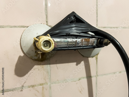 Provisionally repaired shower holder, wrapped with black insulating tape, on an old, pink, dirty tile wall, showing improvisation and negligence