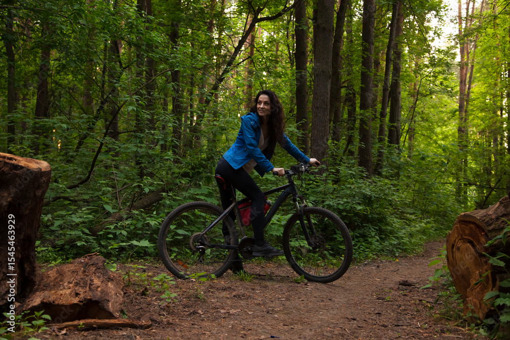 Naklejka premium Beauty smiling sport free woman biking on bicycle on path with logs in green woods in a summer forest. Strong body, legs girl outdoors. Female walking in park in sporty mountain bike.