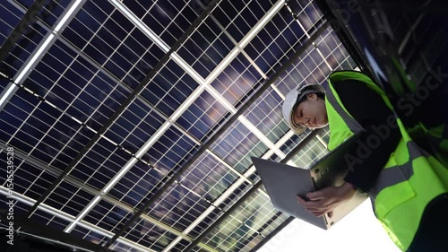 Female engineer inspecting solar panels with a laptop under photovoltaic structure, concept of green energy and renewable technology.