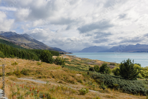 A view of turquoise waters of Lake Pukaki stretching towards hazy mountains under an overcast sky, with dry, golden hills and patches of green vegetation. 