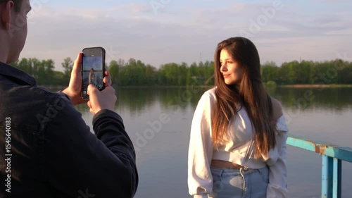 A male photographer takes pictures on his phone of a beautiful long-haired model who stands against the background of a summer landscape
