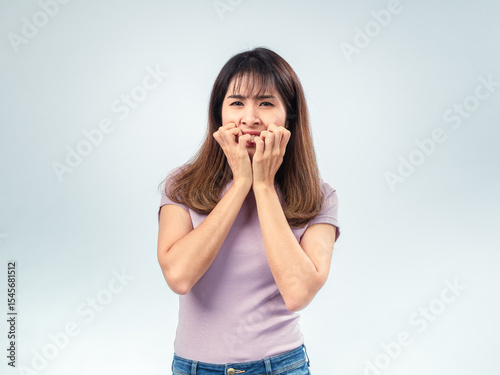 Asian woman in a light purple shirt looking anxious and biting her nails with hands close to her mouth, expressing nervousness against a plain studio background