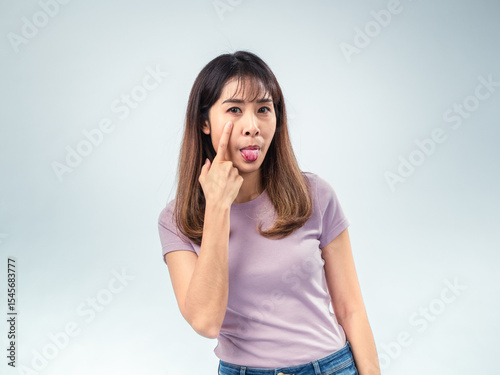 Asian woman making a playful face by sticking out her tongue and pointing to her cheek while wearing a light purple t-shirt, posing humorously in front of a plain light background