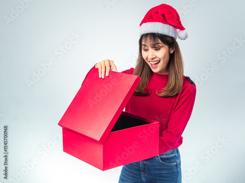 Asian woman in red sweater and Santa hat looking surprised while opening a large red gift box, with joyful expression and festive spirit on a plain light background