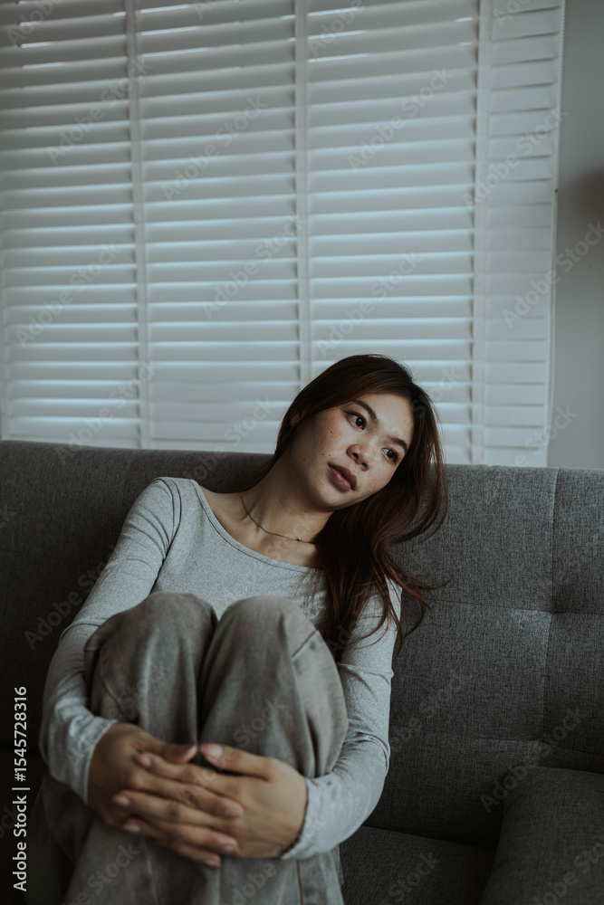 A young woman sits alone on a couch, holding her head in distress. The mood is somber, suggesting stress, anxiety, or emotional exhaustion at home. A young woman sits alone on a couch, holding her head in distress. The mood is somber, suggesting stress, anxiety, or emotional exhaustion at home.