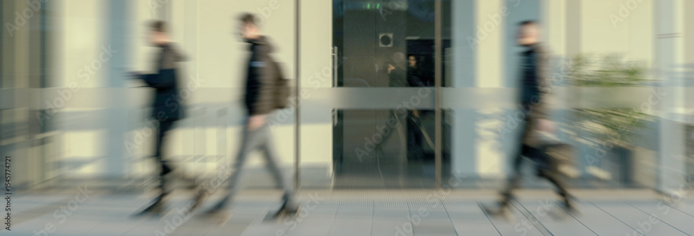 Obraz premium Blurred figures of people are seen walking past glass doors at the entrance of a modern building, conveying a sense of movement and urban activity.
