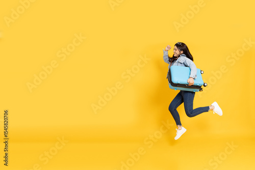 Happy Young Asian traveler woman jumping with blue suitcase bag isolated on yellow background, Tourist in holidays vacation concept