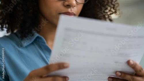 A young businesswoman with curly hair and glasses is seriously reviewing a document at her office desk