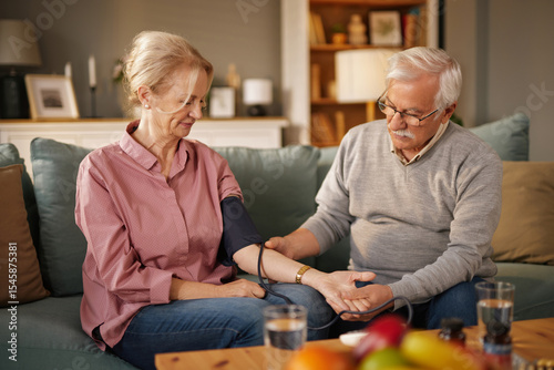 An older man in glasses measures the blood pressure of his smiling wife, who is wearing a pink shirt and jeans, while they sit on their couch in the living room during the daytime.