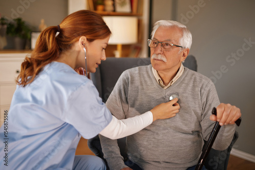 Young female home care nurse uses a stethoscope to examine elderly man with glasses and a walking stick, sitting in an armchair, in his Living Room.