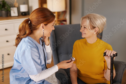 In a home living room, a young female nurse checks an older woman's heartbeat using a stethoscope. The senior woman sits in a chair and holds a cane, smiling at the caring nurse.