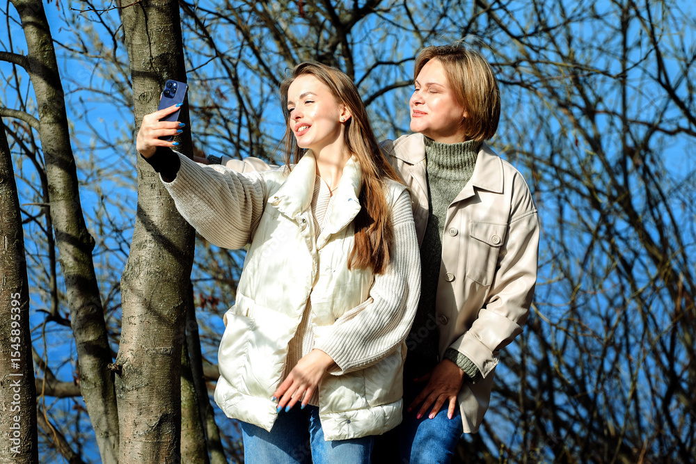 Fototapeta premium Mature mother and adult daughter are doing selfie by mobile phone in summer park, near lake.