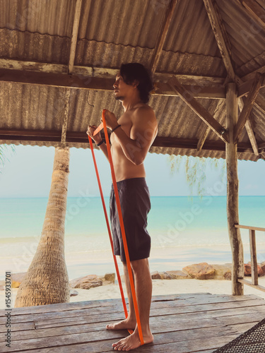 Man exercise and workout with rubber band in the hut by the sea. Beach relaxed vibe.