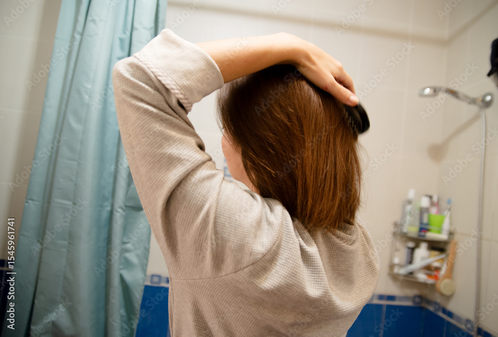 Obraz premium Person seen from behind brushing brown hair in bathroom. Daily self-care and hygiene routine. Features beige sweater, blue curtain, toiletries. Focus on hair care inside.