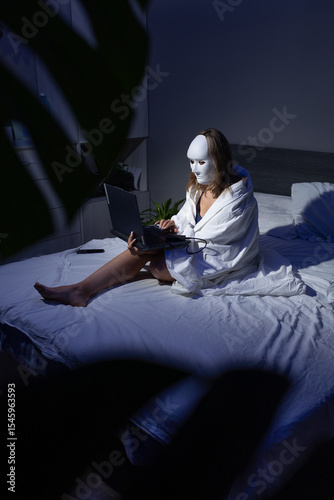 Картината върху платно In a dimly lit room, a person draped in a blanket sits on a bed, using a laptop