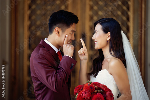 Playful Asian wedding couple smiling and pointing at each other. Bride in white dress, groom in burgundy suit with red bouquet. Happy moment outdoors.