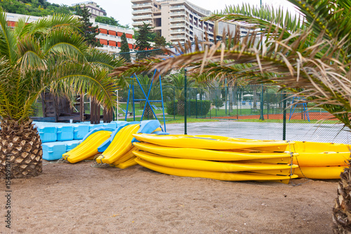 Fototapeta Naklejka Na Ścianę i Meble -  Stack of yellow kayaks on sandy beach with palm trees and resort buildings. Outdoor recreation, coastal leisure and water sports equipment.