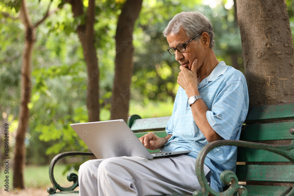 Fototapeta premium Indian senior man working laptop in park. old man wearing blue t-shirts and eye wear glasses while sitting on the bench at the outdoor garden..