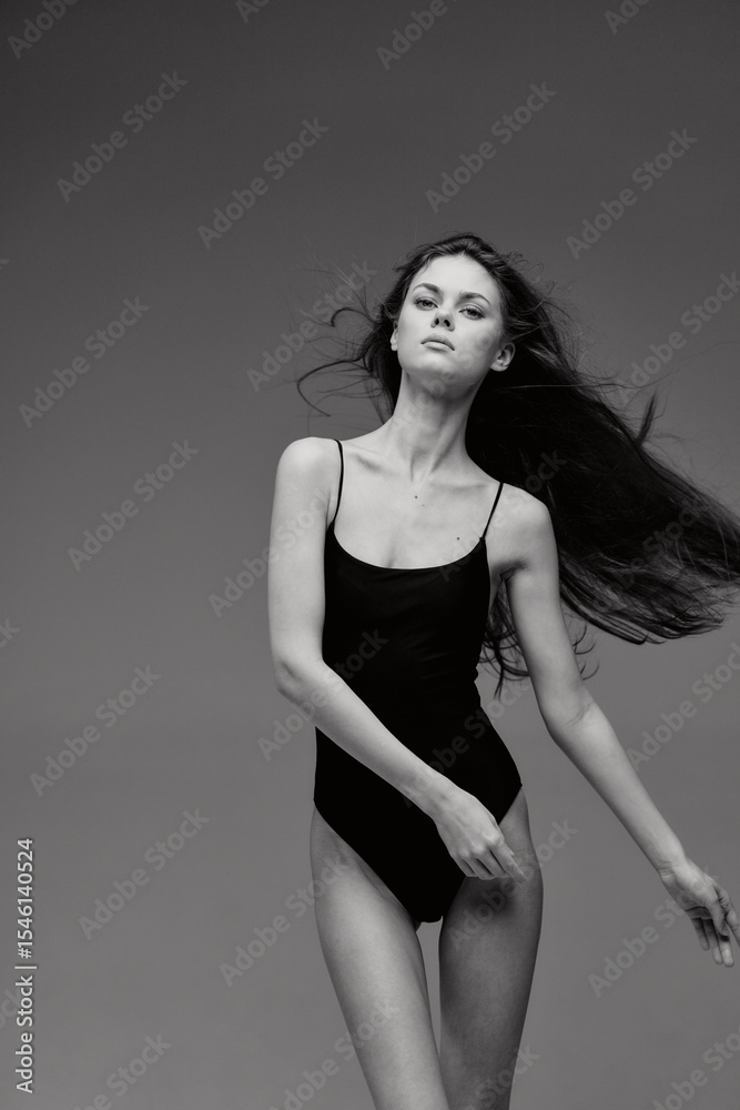 Fototapeta premium black and white woman wearing a black bodysuit poses confidently with flowing hair against a plain gray background, showcasing elegance and minimalistic style.