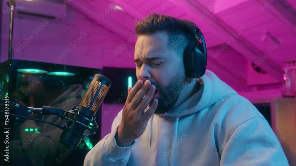 Sleepy Caucasian man guy male gamer streamer yawning slapping face to stay awake playing game with headphones microphone in neon room tired fighting sleep during night gaming session exhausted player