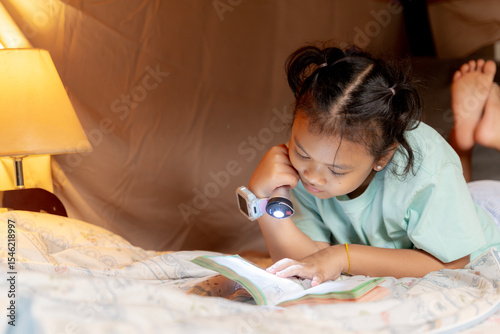 Smiling young girl enjoying a book while lying inside her homemade blanket fort under the table.