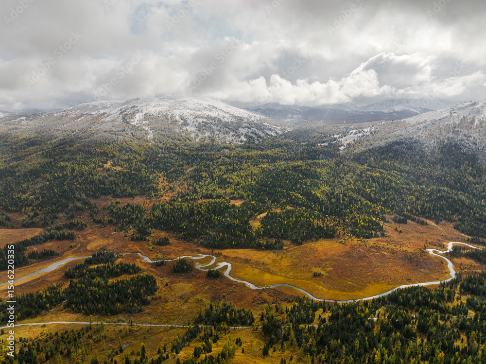 Obraz premium An aerial view depicts a winding river flowing through golden-brown marshland and green forests, with snow-covered mountains in the distance