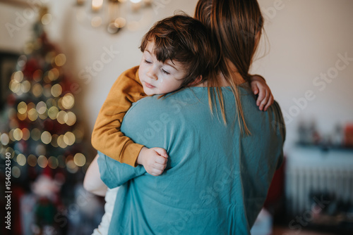 Close-up of a motherly figure affectionately holding a child as a decorated Christmas tree glimmers in soft focus, evoking warmth and the spirit of family during the holiday season.