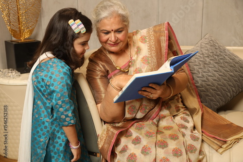 Indian happy grand mother reading story book with her grand daughter in home.