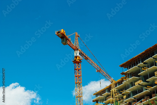 Construction crane operates at building site under clear blue sky with fluffy clouds