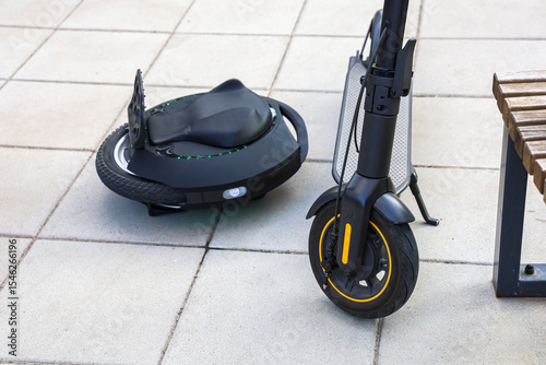 Electric scooter and unicycle parked on a city sidewalk near a wooden bench in bright daylight