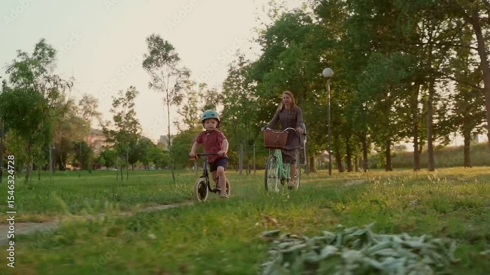 A joyful boy wearing a helmet rides a bicycle with his mom in a sunny park during sunset, enjoying outdoor family time and playful bonding. Slow motion A joyful boy wearing a helmet rides a bicycle with his mom in a sunny park during sunset, enjoying outdoor family time and playful bonding. Slow motion