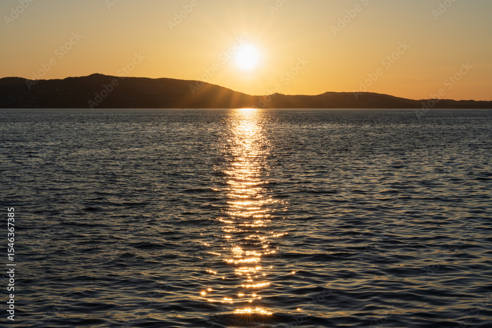 Naklejka premium A picturesque evening landscape in Norway. The bright sun over the hills and its golden reflection on the dark water of a Scandinavian fjord.