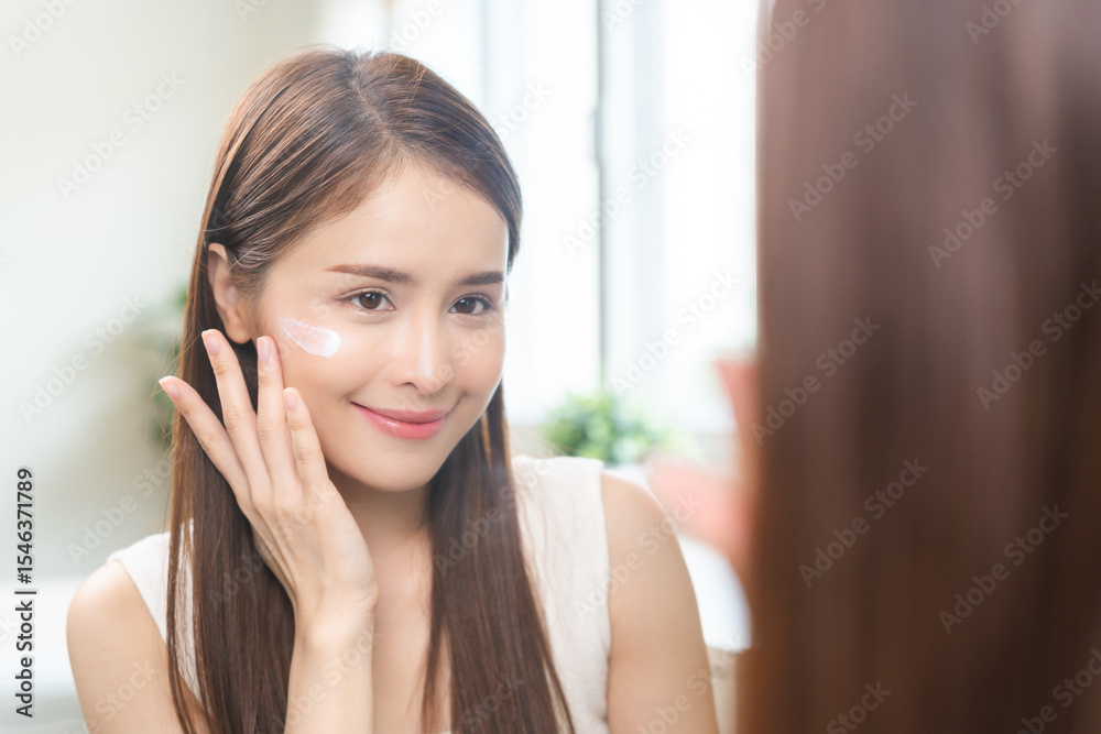 Fototapeta premium Facial beauty skin health care, smile of cute asian young woman looking at mirror, hand applying moisturizer lotion on her face, female putting cream treatment before makeup cosmetic routine at home.