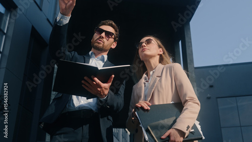 Two stylish teammates businessman talking conversation with businesswoman in sunglasses colleagues businesspeople discuss work documents business man woman with papers pointing city building teamwork