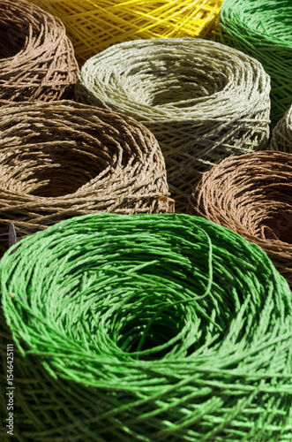 Natural raffia fibers are wound into spools. Skeins are beige, brown, green and yellow. The texture of each spool is a circular pattern.
