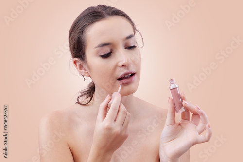 Young woman portrait applying lipstick with lip brush applicator holding lipstick bottle near her face and looking at it. Natural skin with smooth foundation. Beauty routine concept. Isolated on beige