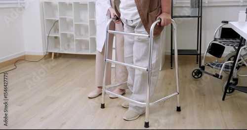 Asian elderly woman expressing pain while trying to stand using walker during slow rehabilitation session in clinic while Caucasian physiotherapist monitors movement and supports painful transition