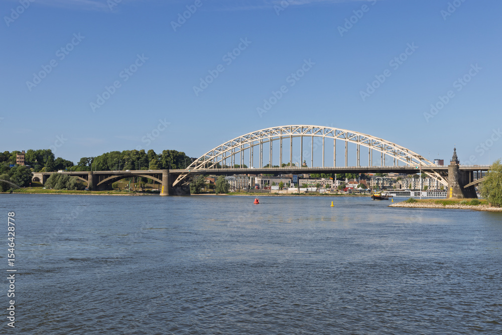 The impressive Waalbrug, a landmark arch bridge, spans the Waal River in Nijmegen under a clear blue sky, connecting the vibrant city with its lush surroundings. The impressive Waalbrug, a landmark arch bridge, spans the Waal River in Nijmegen under a clear blue sky, connecting the vibrant city with its lush surroundings.