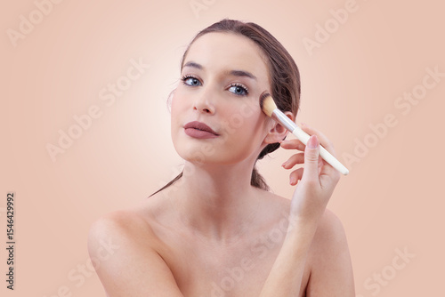 Portrait of a smiling young woman applying blush with a makeup brush while looking into the camera with her blue eyes. Clean, minimal beauty routine concept. Isolated on beige background.