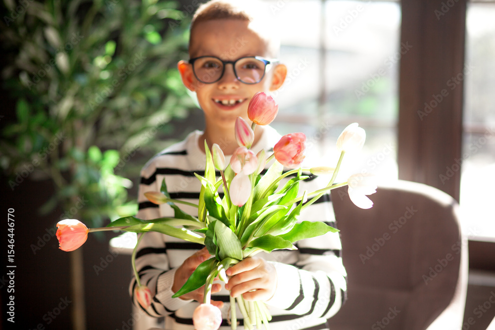 Fototapeta premium A 7-year-old child in glasses and dresses striped vest, poses at the school table indoor. The boy holds flowers