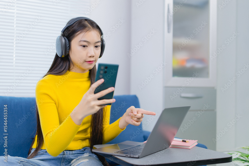 Asian freelancer wearing headphones working remotely comparing information on her laptop and smartphone while sitting on a blue sofa in her living room Asian freelancer wearing headphones working remotely comparing information on her laptop and smartphone while sitting on a blue sofa in her living room